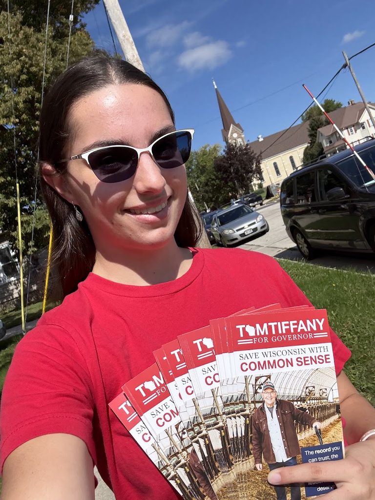 Volunteer in a red shirt and sunglasses holds several Tom Tiffany campaign brochures outdoors.