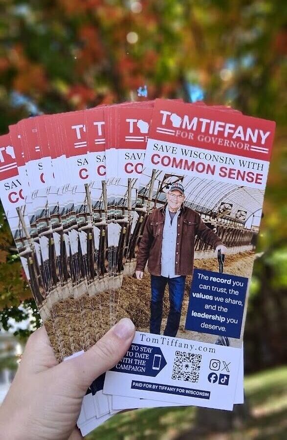 A hand holds a stack of Tom Tiffany for Governor campaign brochures in front of autumn trees.