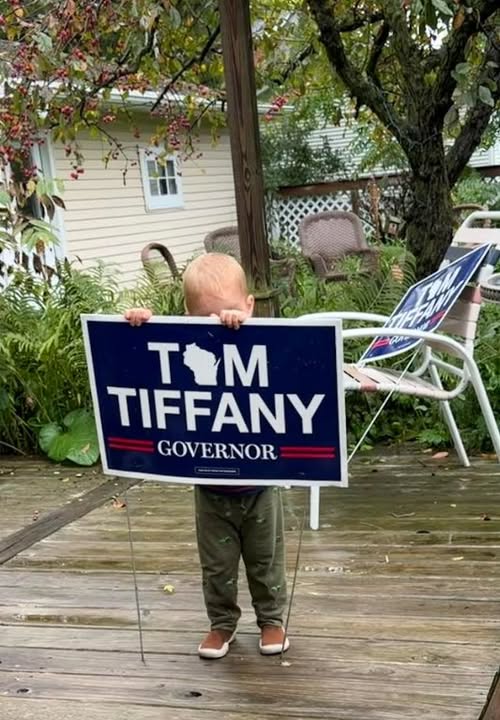 Young child holding a Tom Tiffany yard sign, accompanying information about RPSC headquarters, yard-sign pickup and delivery, office hours, and volunteer opportunities.